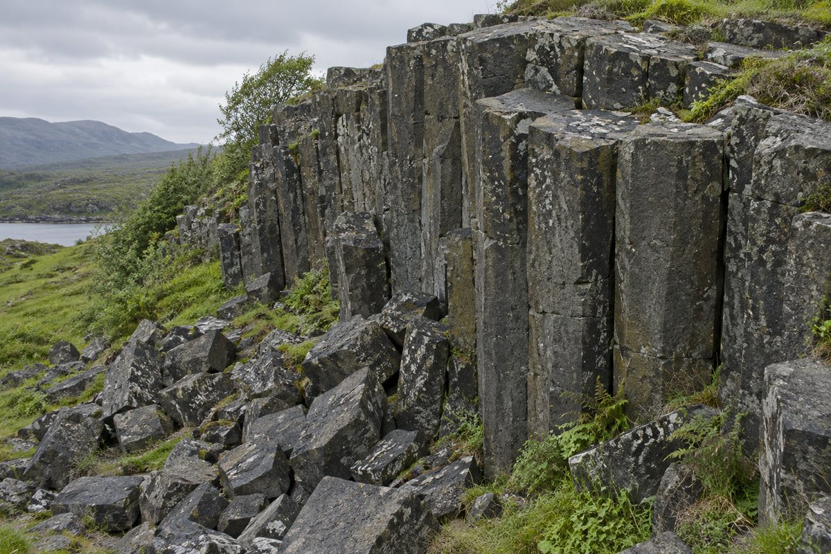 Dark polygonal rock columns rise above a pile of broken angular fragments in a green landscape.