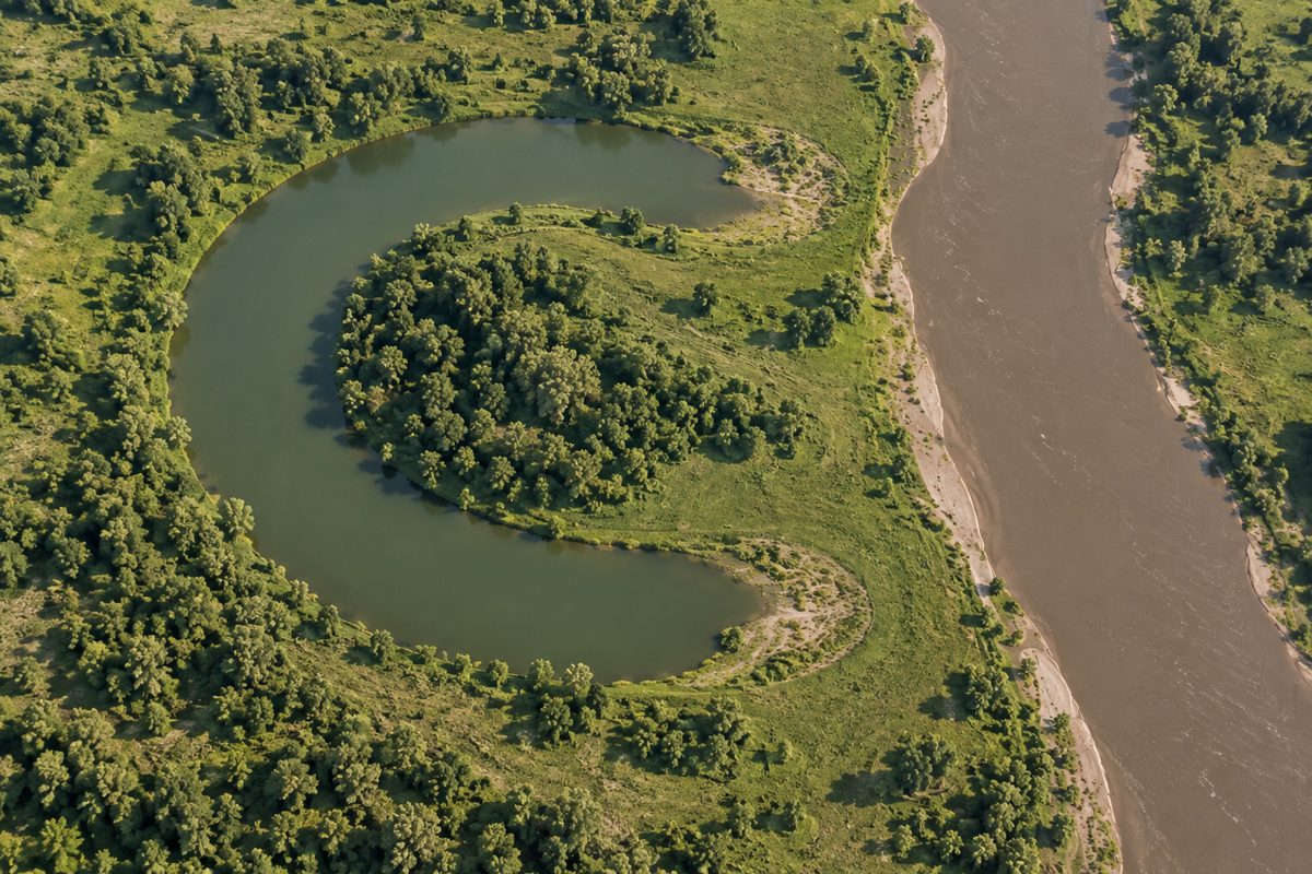 A curved lake lies beside a muddy river, with grassy ground separating both ends of the lake from the channel.